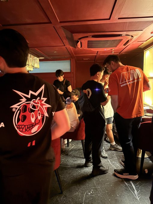 A group of young people gather in a restaurant or lounge with red lighting and coffered ceilings, with one person in the foreground wearing a black shirt featuring a cartoon strawberry graphic.