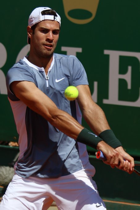 A male tennis player wearing a blue Nike shirt and white cap executes a two-handed backhand swing as he makes contact with a yellow tennis ball.