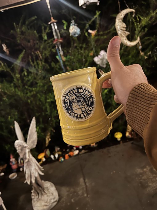 A hand holds a yellow 'Death Wish Coffee Co.' mug at night, with garden decorations including a moon ornament and angel statue visible in the background.