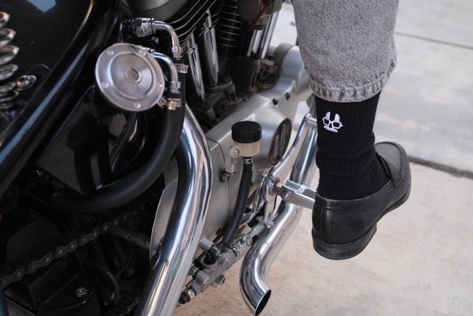 A rider's foot, clad in a black shoe and black sock featuring a white skull and crossbones logo, rests on the footpeg of a chrome-accented motorcycle, ready to rev up in response to the "Vroom?" call.