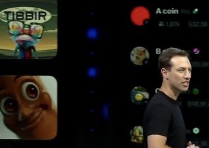 The image features a man standing next to a computer screen, with several cartoon-like images displayed behind him. These images include a character from a game called "Cool @base update $tibbir", as well as other cartoon-style images. The man appears to be giving a presentation or speech on stage, and the image captures this moment in time.