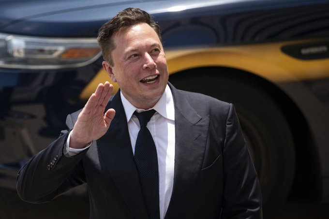 Elon Musk in a dark suit and tie waving, standing in front of a black SUV.
