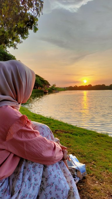 A person wearing a pink hijab and a floral skirt sits on the grass, looking out over a calm body of water as the sun sets.