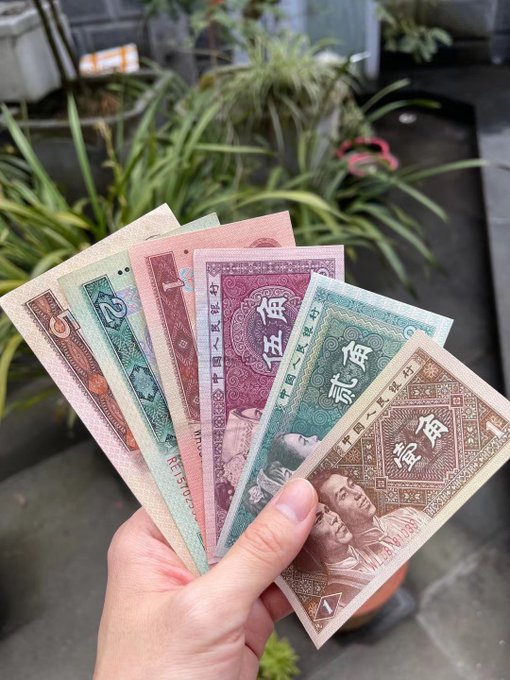 A hand fans out several Chinese yuan banknotes in denominations of 1, 2, and 5, displayed against a backdrop of green plants.