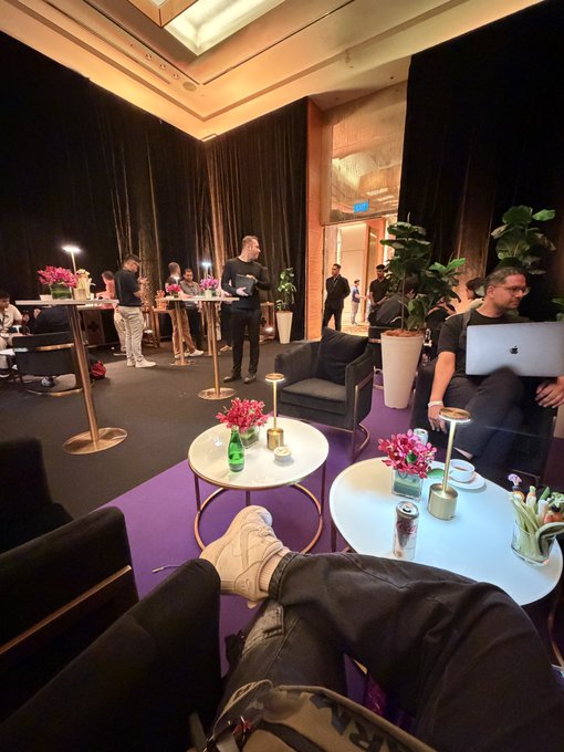 A first-person perspective from someone relaxing with their feet up in a lounge area, showing a purple carpet, round tables with flowers, and other people working on laptops in the background.