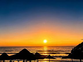 A vibrant sunset silhouettes beach umbrellas and rocks along the shoreline as the sun dips toward the ocean horizon.