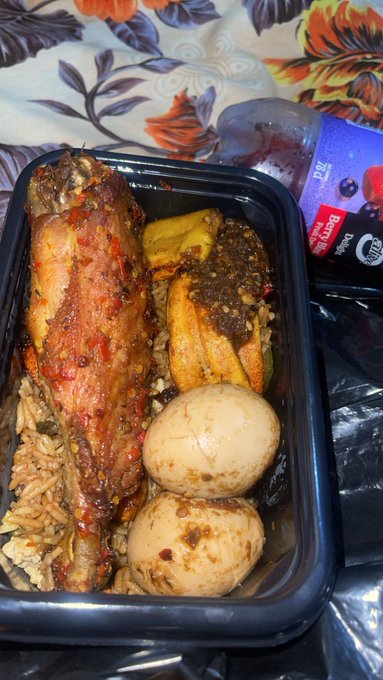 A black takeout container holds a meal of rice, jerk-seasoned meat, plantains, and two whole boiled eggs, with a bottle of berry-flavored drink beside it.