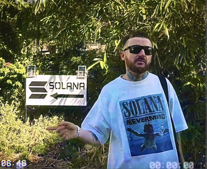 A man wearing sunglasses and a white t-shirt with 'SOLANA NEVERMIND' graphic stands outdoors pointing at a Solana directional sign, with solar panels visible on the sign and a timestamp '06:46' in the corner.