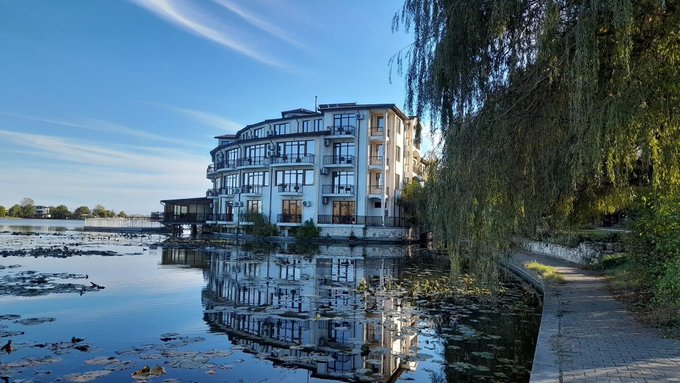 A scenic view of a multi-story building next to a lake reflecting the blue sky, with a paved walkway lined by trees.
