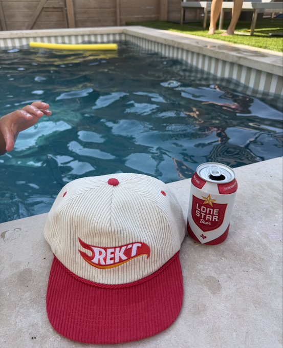 A cream and red corduroy baseball cap with a 'REKT' logo sits on a poolside ledge next to a can of Lone Star Beer, with a swimming pool visible in the background.