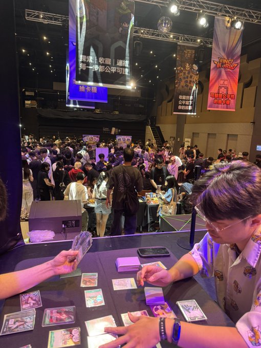 A crowded convention hall scene shows people playing a Pokémon trading card game, with banners in Chinese and Japanese including 'VINE 6/17' visible.