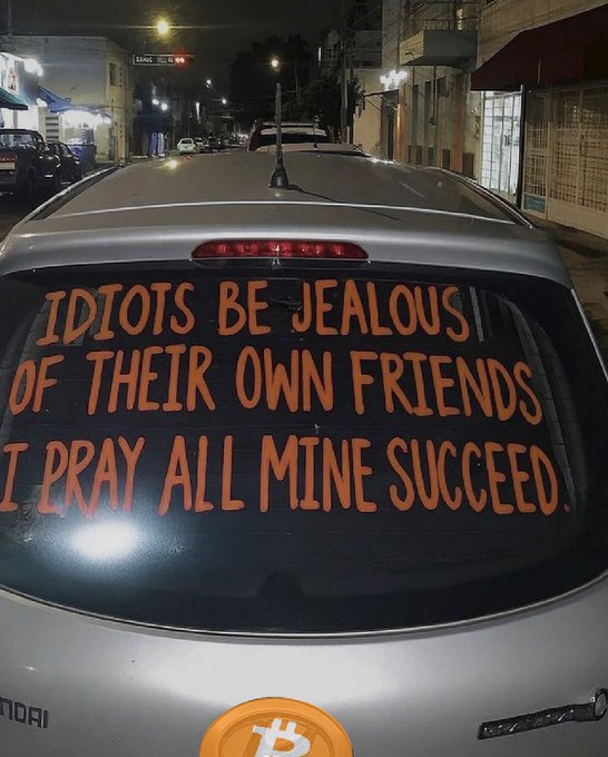 The back window of a silver Honda car at night displays hand-painted orange text reading 'IDIOTS BE JEALOUS OF THEIR OWN FRIENDS I PRAY ALL MINE SUCCEED.' alongside a Bitcoin logo sticker.