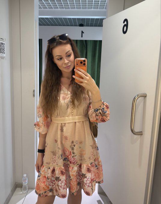 A mirror selfie of a woman with long dark hair wearing sunglasses on her head and a floral-patterned dress, holding an orange phone case in what appears to be a clothing store fitting room.