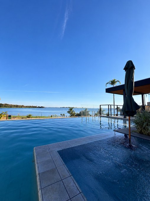 A luxurious infinity pool overlooks a serene ocean view under a clear blue sky, with a closed umbrella and modern resort architecture on the right. The image's tranquil, high-end setting complements the "GOOD MORNING" post, hinting at a leisurely lifestyle often associated with crypto prosperity and inviting $SOL address drops.