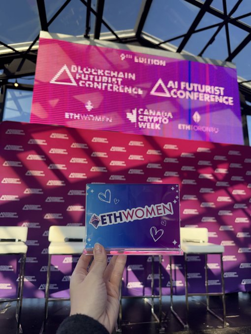 A photo taken at an event showing a hand holding an 'ETHWOMEN' placard in front of a stage backdrop advertising the 'Blockchain Futurist Conference 2nd Edition' and 'AI Futurist Conference' with logos for ETHWomen, Canada Crypto Week, and ETHToronto.