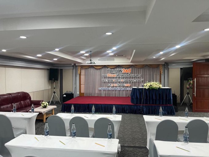 An empty seminar room is set up with a stage displaying a Bitcoin 360 AI banner, rows of tables with water bottles and notepads, and floral arrangements on the stage.