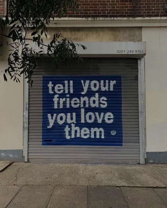 A blue mural painted on a metal roller shutter displays the text 'tell your friends you love them.' in white lettering, with a phone number '0207 249 9703' visible on the wall above.