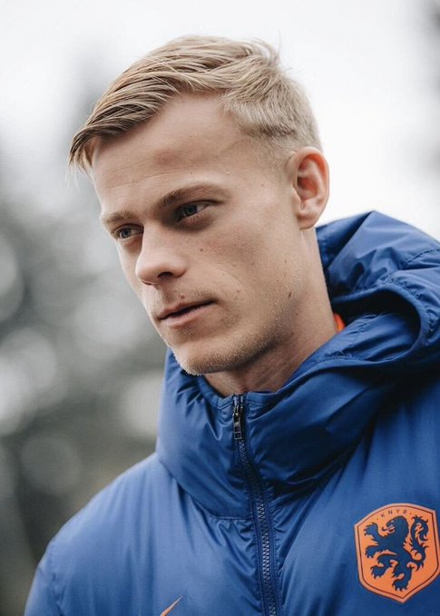 A portrait of a blonde man wearing a blue jacket featuring the orange crest of the Royal Netherlands Football Association (KNVB).