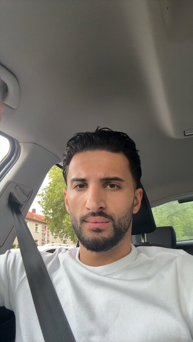 A selfie of a bearded man in a white t-shirt sitting in the driver's seat of a car.
