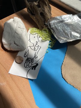 An overhead shot of an artist's workspace showing a black ink sketch of a figure on white paper, surrounded by crumpled tissue, a bronze coin, pieces of driftwood, aluminum foil, and bright blue paper on a wooden surface.