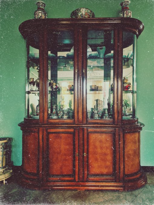 A tall, ornate antique wooden display cabinet with a curved glass center section, showcasing various ceramics and decorative items. The lower half features wood panel doors with a burl wood finish, set against a green wall.