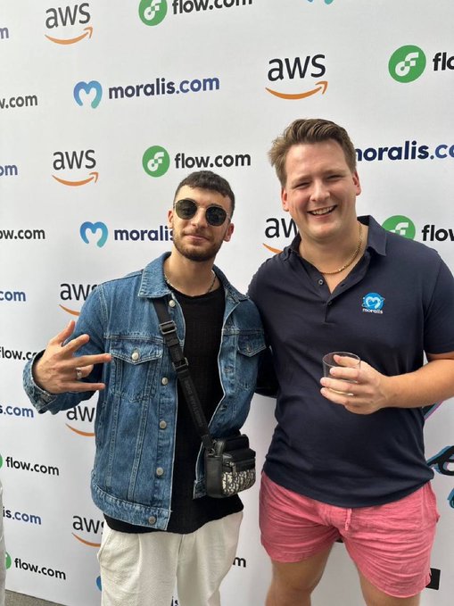 Two men posing together at an event with sponsor logos (aws, moralis.com, flow.com) in the background; one wears a denim jacket and sunglasses, the other a navy polo with a Moralis logo and pink shorts.