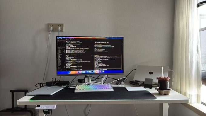 A minimalist home office desk setup featuring a large monitor displaying code, a white RGB mechanical keyboard, a laptop, and a boba drink beside a window.