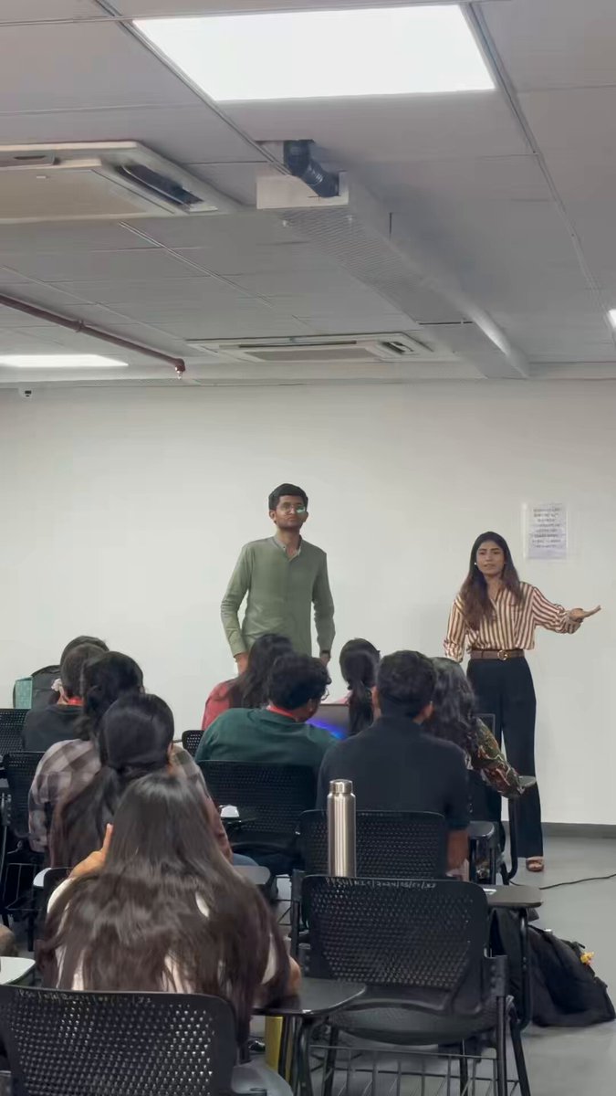 The image shows a classroom setting with two people standing at the front of the room, one person wearing a green shirt and another person wearing a striped shirt. They are giving a presentation or lecture to an audience consisting of students seated in chairs around them. The students appear engaged and attentive as they listen to the speakers.