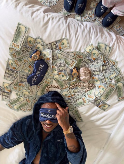 A person in a dark robe and 'Madstash Society' eye mask lies on a bed covered with scattered $100 bills, with a copper-colored bust and other slippers visible around them.