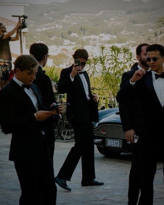 Several men in black tuxedos and sunglasses on a cobblestone street near a classic Aston Martin, with one filming on a camera.