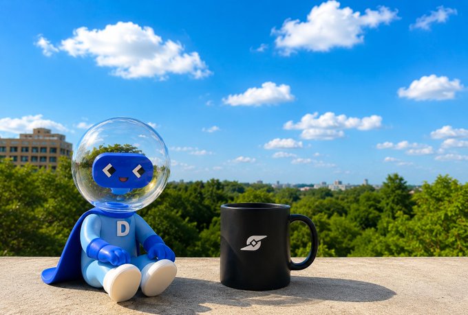 The image features a blue toy robot with a helmet sitting next to a black coffee mug. The robot has a blue shield, which matches its overall color scheme. The mug also has a blue design on it. Both items are placed outdoors, possibly on a ledge or table. The background shows a beautiful view of trees and buildings under a clear sky, creating an interesting contrast between the man-made objects and their natural surroundings.