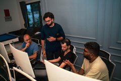 Four people gathered around computer monitors in what appears to be an office or gaming setup, with one man standing behind and three seated at the desks.