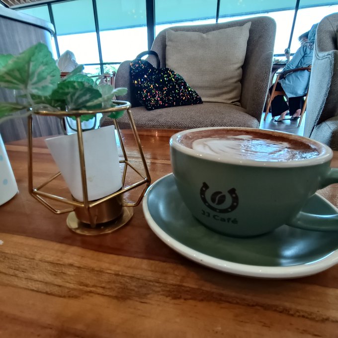 A matcha latte in a teal cup with the '33 Café' logo served on a saucer at a wooden table next to a small potted plant, with a chair visible in the background