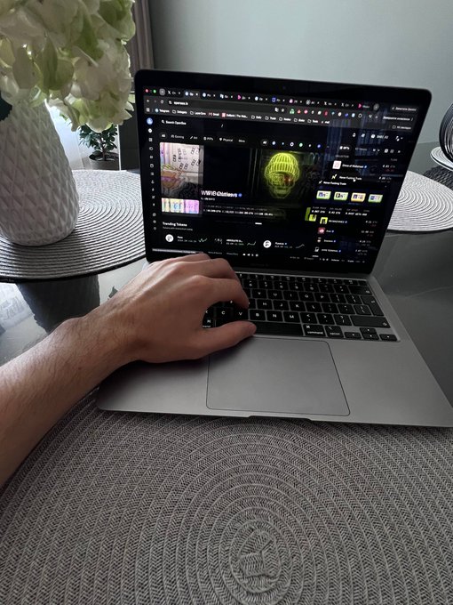 A first-person view of a hand navigating Spotify on a MacBook laptop on a dining table, with a vase of white flowers in the background.