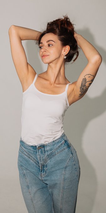 A young woman with her hair in a bun poses with both hands behind her head, wearing a white tank top and blue jeans, with a tattoo visible on her left upper arm.