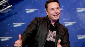 The image features a man wearing a black jacket, standing against a blue background that includes logos for Twitter and Tesla. The man has his arms outstretched with two thumbs up signs, indicating positivity or agreement. The text "Elon Musk" is also visible in the image. This could be part of an advertisement or promotional material related to Elon Musk's company, Tesla.
