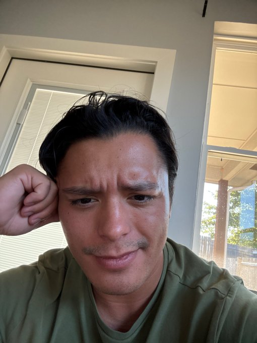 A close-up selfie of an Asian man in an olive green shirt looking at the camera with a concerned expression, resting his head on his hand.
