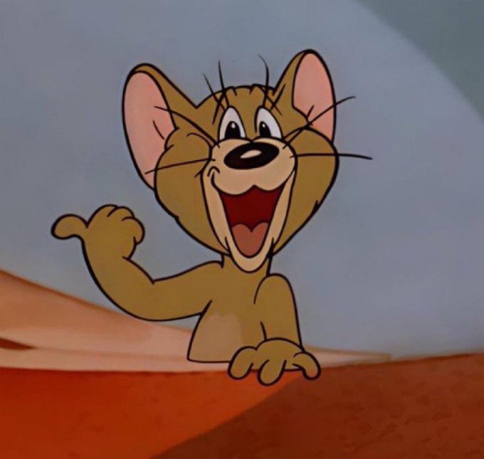 A cartoon character, identified as Tom from "Tom and Jerry," stands on a red carpet with arms outstretched, wearing a yellow shirt and blue pants against a light blue sky backdrop.