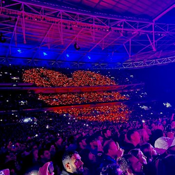 A concert venue interior illuminated with blue lighting, featuring a large heart-shaped display of red lights suspended above a crowd holding pink balloons.