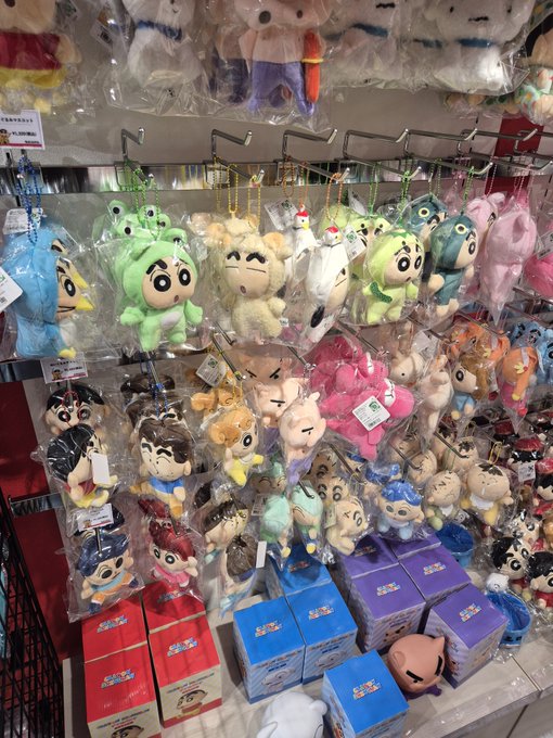 A retail display rack filled with colorful plush toys in plastic packaging, including characters resembling Crayon Shin-chan, and boxed figures at the bottom.