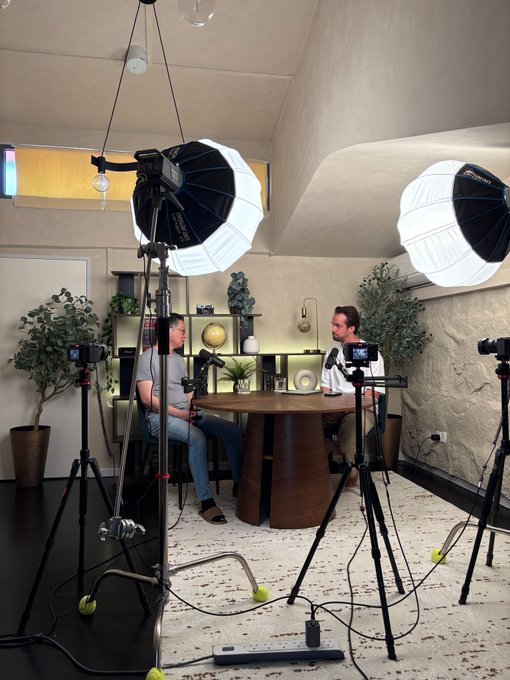 Behind-the-scenes shot of a podcast setup featuring two people seated at a round table with multiple cameras on tripods and professional lighting equipment in a decorated studio.