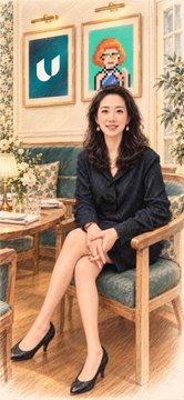 A woman in a black blouse and heels sits smiling on a green chair in a restaurant, with framed artworks of a whale and a cartoon figure hanging behind her, fitting the post's scene of enjoying a stylish, detail-oriented Michelin-level dining experience in Singapore.