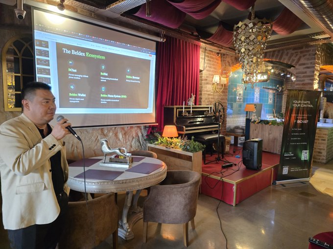 The image shows a man standing behind a microphone, likely giving a speech or presentation at an event. The room has a stage with a piano and a projector screen displaying information about the event. There are also chairs arranged around the stage for attendees. A sign is visible in the background that reads "IBW 2026 sonrasında @BeldexCoin tarafından düzenlenen etkinliğe katılım Açık konuşmak gerekirse klasik bir networking etkinliğinden çok daha samimi bir ortam var Türk mutfağının izmirleri". The man is wearing a suit, and the room has a red curtain.