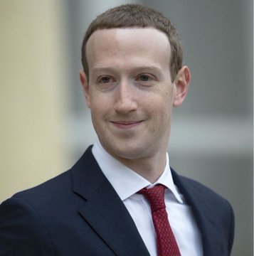 A man in a dark suit, white shirt, and red tie smiles slightly at the camera in an outdoor portrait.