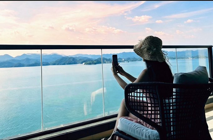 A woman wearing a sun hat sits on a balcony overlooking a serene ocean and mountains at sunset, checking her phone while enjoying the view. The image perfectly captures the post's message of crypto-enabled location independence, showing how digital traders can monitor markets from a luxurious, scenic destination rather than a traditional office.