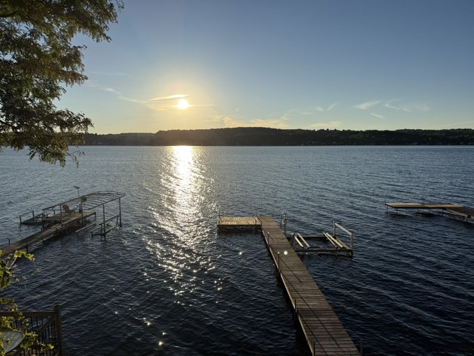 A serene photograph of a wooden dock extending into a calm lake, with the setting or rising sun reflecting brightly on the water.
