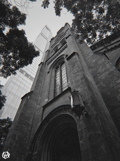 A black and white low-angle photograph of a Gothic-style stone church or building with a pointed arch entrance, framed by trees and a modern skyscraper in the background.