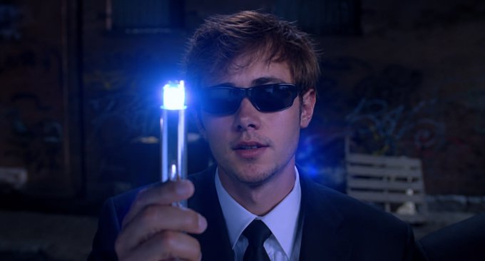 A man with tousled hair and dark sunglasses in a suit holds up a glowing blue cylindrical object, illuminated against a dark, graffiti-covered industrial background.