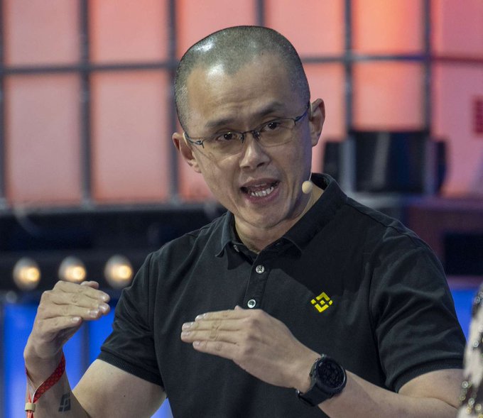 Binance founder Changpeng Zhao (CZ), wearing a black polo with the yellow Binance logo, speaks animatedly while gesturing with both hands during what appears to be a public talk or interview. The image illustrates his ongoing criticism of traditional banking, as he advocates for cryptocurrency as a more trustworthy alternative.
