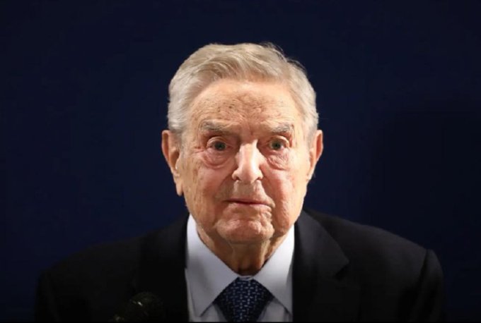 A portrait of an elderly man with gray hair, wearing a dark suit and blue tie, photographed against a dark blue background.